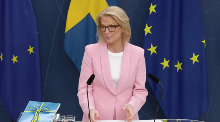 En kvinna i rosa kavaj framför svenska flaggan och EU-flaggan.