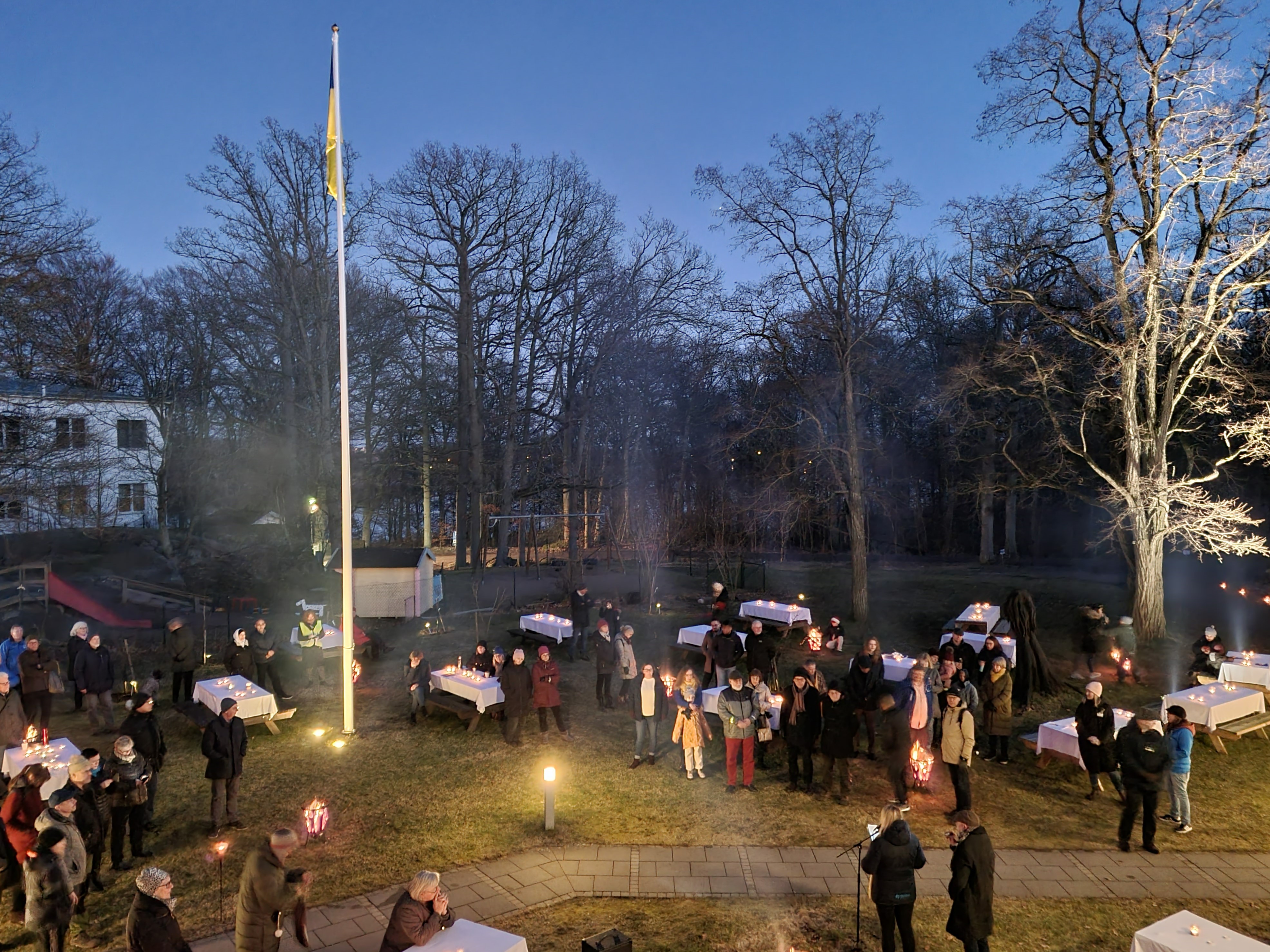 Mörk utomhusmiljö, ovanifrån. Många människor står kring bord med vita dukar.