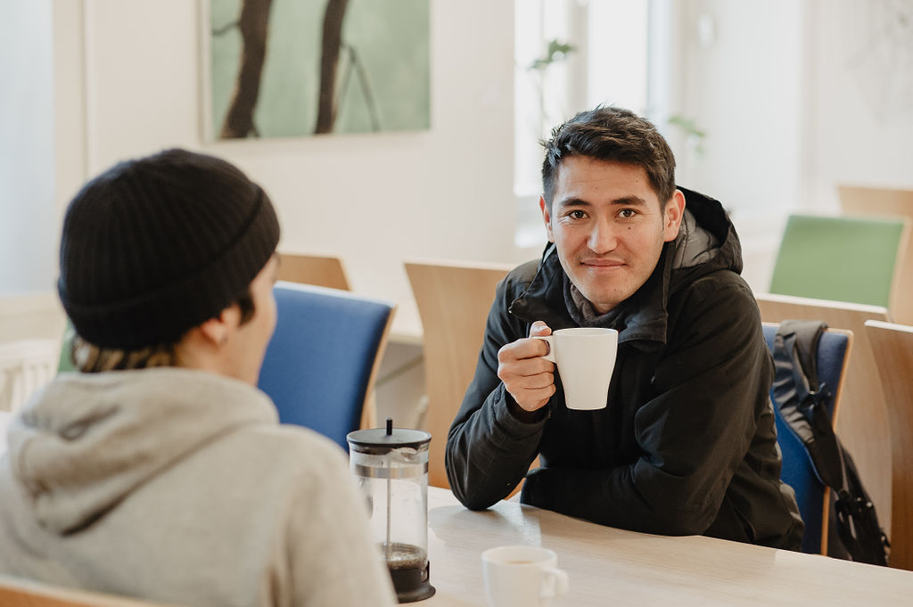 Två personer sitter vid ett vitt bord. En med ryggen till. Den andra håller en vit kaffekopp, ler.