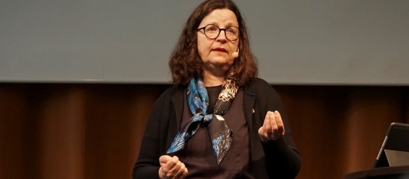 Kvinna med runda glasögon står på en scen och talat.