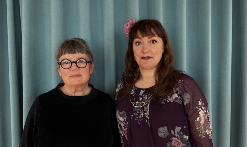 Två kvinnor tittar in i kameran med ett blått draperi i bakgrunden. 