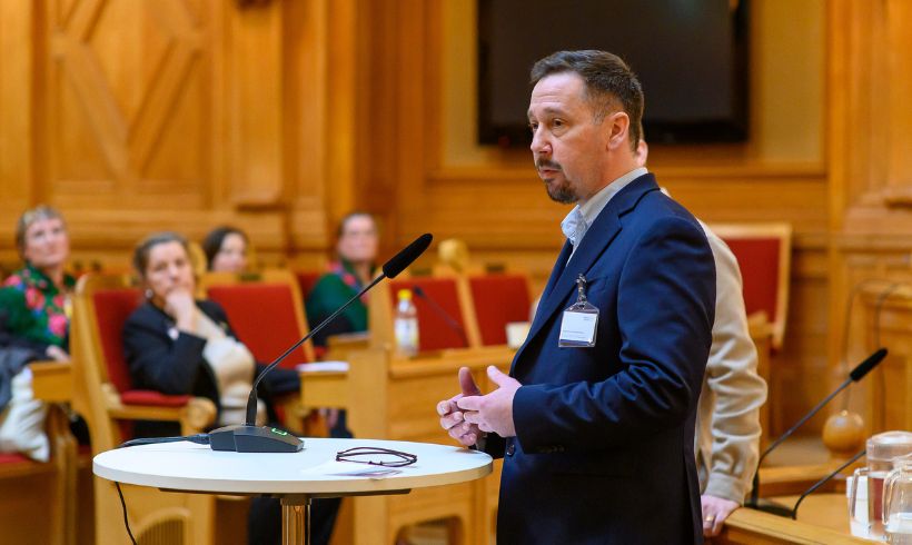 En man står i en talarstol framför fyra personer. 